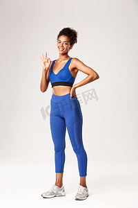 全长的非洲裔美国女运动员身体健壮，表现出赞同的好姿态，推荐健身房或锻炼计划，站在白色背景上感到满意