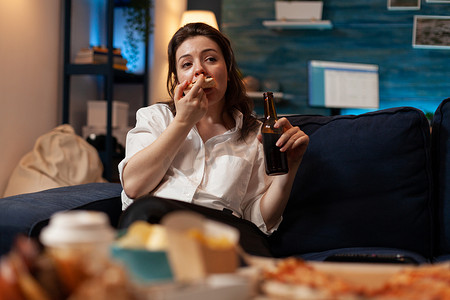坐在沙发上拿着啤酒瓶看电视的女人正在吃一片热披萨外卖