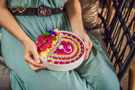 年轻女子坐在沙发上吃地中海早餐，吃健康的热带早餐、热带水果冰沙碗，装饰着彩色酸奶、姜黄和螺旋藻的图案。