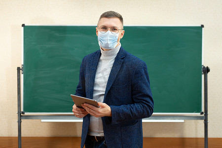 科创教室摄影照片_一名戴着医用口罩的男老师站在教室黑板前，手里拿着平板电脑。
