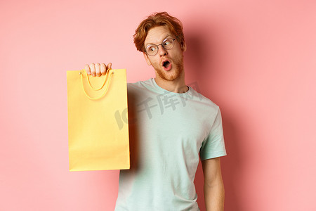 黄色活动促销摄影照片_红头发、戴着眼镜和 T 恤、拿着并看着黄色购物袋、在促销活动中购买礼物、站在粉红色背景上的风趣帅哥