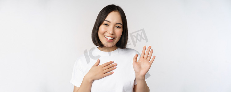 美丽的韩国女孩举手的画像，自我介绍，手放在心上，问候，站在白色背景上