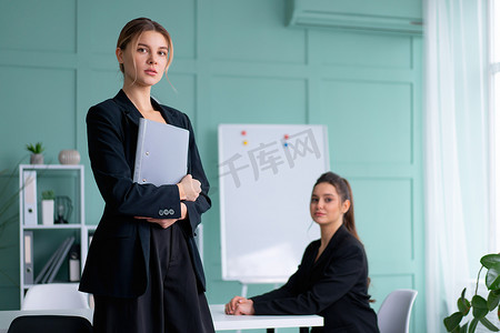 两位年轻女性领导人在办公室穿着黑色西装，一位女士拿着文件夹站着，另一位女士坐在桌子上看着相机。