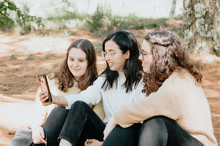 智能社区摄影照片_三名年轻女学生在阳光明媚的日子里在森林里的手机里找东西，友谊概念，爱情