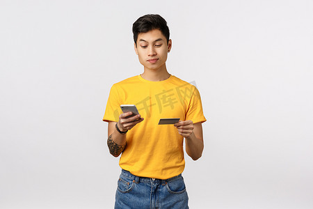 身穿黄色 T 恤、手持智能手机和信用卡、输入数字号码在线购买、购物互联网、支付申请费用、站立白色背景的英俊亚洲年轻人