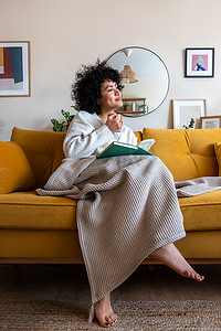 沉思快乐的非洲裔美国女人坐在沙发上放松地喝着咖啡，在家看书。