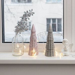 灰色和粉红色的陶瓷圣诞树、带有人造银树枝的玻璃瓶和花环装饰着圣诞节的窗台。