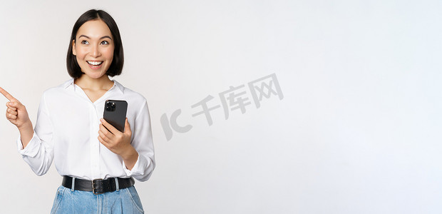 手机版k线摄影照片_兴奋的亚洲女孩在移动应用程序上购物，拿着手机，手指留在空白的复制空间，显示促销文本，站在白色背景下的智能手机