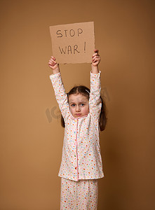 军事冲突摄影照片_可爱的心烦意乱的孩子，穿着睡衣的悲伤欧洲小女孩拿着停止战争社交信息纸板海报，站在米色背景上，上面有广告复制空间