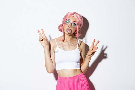 假发宣传摄影照片_戴着粉红色假发的漂亮女孩吹口香糖，脸上冒泡泡，展示和平标志，傻傻地站在白色背景上