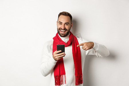 满意的留着胡须的男人用手指指着智能手机，在手机上查看互联网促销优惠，站在白色背景上