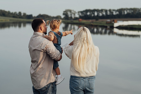 站在户外的年轻家庭的后视图，父亲手里拿着他的孩子，小女儿向她的爸爸和妈妈展示湖