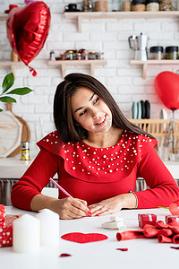 气球情书摄影照片_身穿红裙的年轻浪漫女人坐在装饰精美的厨房里写情书