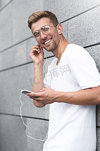 快乐的家伙穿着白色 T 恤在街上，用耳机听音乐，拿着手机。