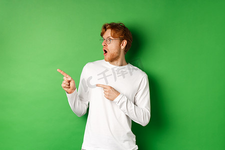 戴着眼镜的红头发男人印象深刻，他看着促销，用惊奇的脸指着左边，站在绿色背景上的长袖 T 恤