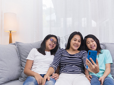 可爱的视频摄影照片_幸福的家庭坐在客厅的沙发上与某人一起在手机上交谈，微笑的母亲通过智能手机与家人交谈显示可爱的小孩视频通话。