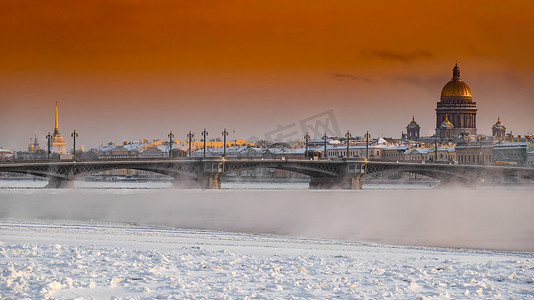 日落时圣彼得堡的冬季全景、艾萨克大教堂和布拉戈维申斯基桥的背景、冰冻涅瓦河上的蒸汽、橙色的天空