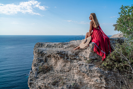 飘逸长裙摄影照片_一个穿着红色长裙、头发飘逸的女孩坐在海上的一块岩石上。