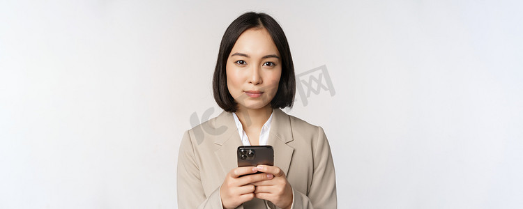 亚洲女商人穿着西装，拿着手机，使用智能手机应用程序，对着镜头微笑，白色背景