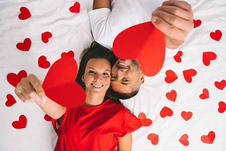 爱心幸福摄影照片_幸福的年轻夫妇相爱，躺在家里的白床上，手里拿着红纸心