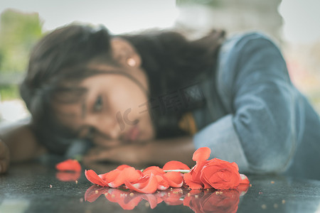 孤寂摄影照片_有选择地关注红玫瑰花瓣，孤独的年轻青少年低着头悲伤地坐在桌子上 — 爱情分手或破碎的心的概念。
