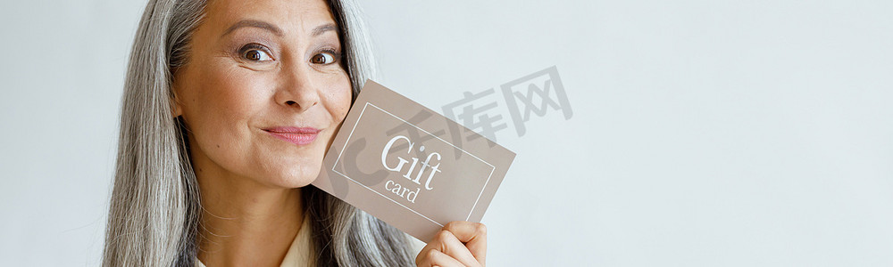 快乐的灰发亚洲女性拿着浅灰色背景的礼品卡