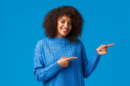 可爱的成年非洲裔美国女性，留着非洲发型，手指向右，建议方向，访问网站或下载应用程序，展示出色的产品，满意地微笑着，蓝色背景