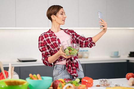 漂亮的家庭主妇穿着格子衬衫，留着鲍勃发型，一边用智能手机自拍或打视频电话，一边烹饪新鲜沙拉。