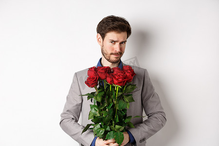 优柔寡断、留着胡须的西装男，手捧玫瑰花，不确定地看着镜头，进行浪漫约会，站在白色背景上