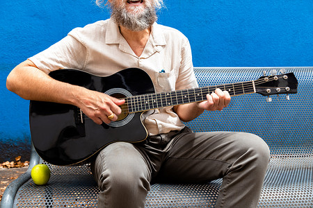 坐弹吉他摄影照片_面目全非的成熟男人，留着胡子，弹吉他，坐在蓝色背景的长凳上唱歌。
