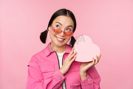 戴墨镜的时尚亚洲女友的形象，猜测礼盒里有什么，心形礼物，站在粉红色背景上