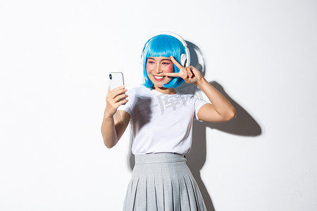 可爱的亚洲女孩打扮成动漫人物的肖像，戴着耳机和蓝色短假发自拍，在智能手机上拍照，站在白色背景上