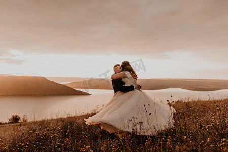 一对恩爱的新婚夫妇穿着白色的裙子和西装拥抱在夏日高高的草地上接吻