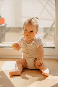 一个善良的 7 个月小女孩穿着紧身连衣裤坐在家里的阳台门附近。