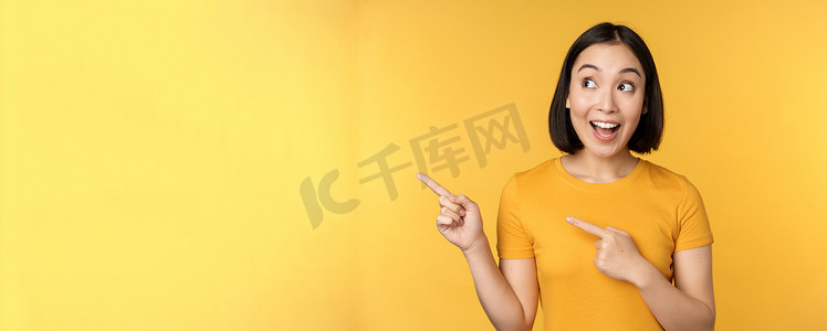 快乐的亚洲女孩的肖像，指着手指，向左看，惊讶地笑着，查看促销横幅，在黄色背景下展示广告