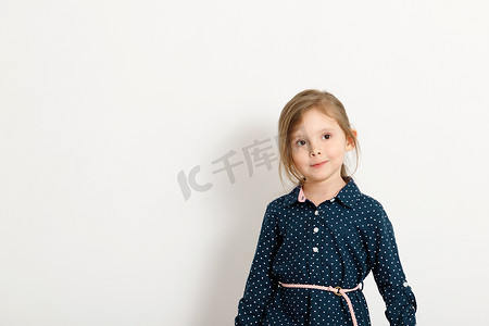 4-6 岁的可爱小女孩穿着一件带圆点的蓝色连衣裙，站在白墙的背景下