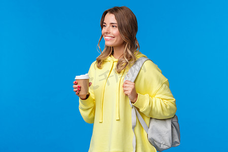 大观念摄影照片_无忧无虑的漂亮年轻女学生在上大学时喝着纸杯咖啡，背着背包，带着愉快轻松的微笑、生活方式和人的观念看着一旁