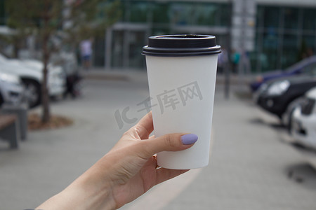 早餐和咖啡主题：一个女人的手拿着一个带有黑色塑料盖的白色纸咖啡杯。