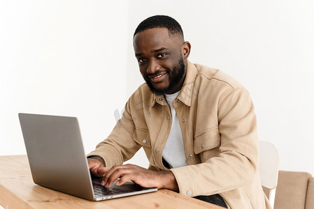坐在家庭办公室的桌子旁，在笔记本电脑上工作时，微笑着看着相机的非洲裔美国人的画像。