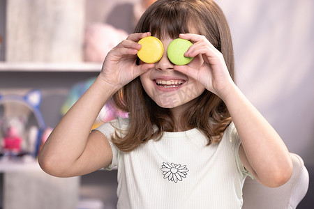 十几岁的女孩玩着甜点马卡龙，把饼干像眼镜一样放在眼睛周围，然后把手扔到两边。