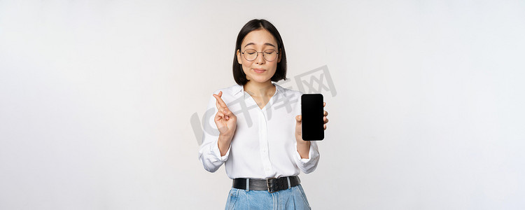 充满希望的亚洲年轻女性，展示手机屏幕、应用程序界面和手指交叉，希望得到 smth，许愿或支付，站在白色背景上