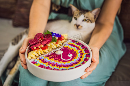 年轻女人坐在沙发上吃地中海早餐，和她的猫一起吃健康的热带早餐，冰沙碗里放着热带水果，上面装饰着五颜六色的酸奶、姜黄和螺旋藻。