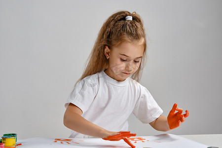 穿着白色 T 恤的小女孩坐在桌子旁，拿着什么和彩色颜料，用手在上面画画。