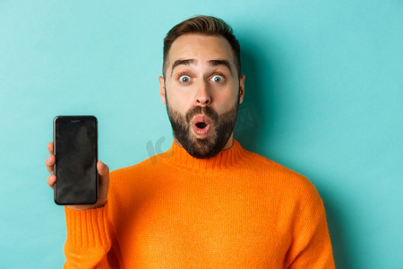 留着胡须的帅哥穿着橙色毛衣的特写，展示智能手机屏幕并微笑，展示在线促销，绿松石色背景