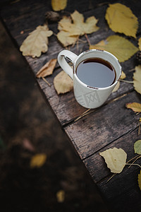秋季公园的一张旧木桌上放着一杯茶和咖啡、散落的黄叶和松果。