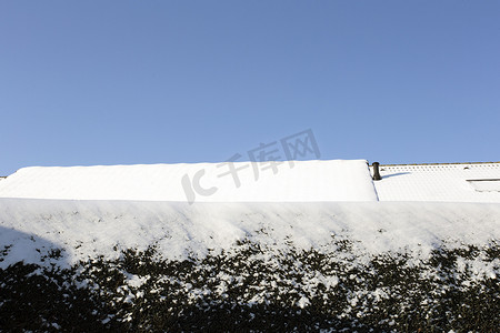雪屋顶摄影照片_冬天覆盖着雪的绿色树篱、明亮的蓝天和屋顶舒适的房屋，复制空间冬季景观