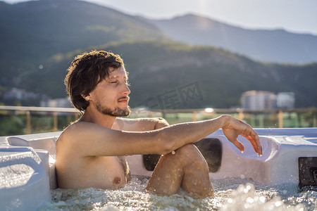 绿色大山背景下享受快乐旅行时光度假生活时，年轻无忧无虑的快乐微笑男子在热水浴缸中放松的肖像