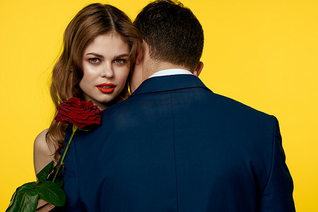 丝质裙子摄影照片_一个浪漫的男人拥抱一个穿着红色裙子的女人，手里拿着一朵黄色背景的玫瑰