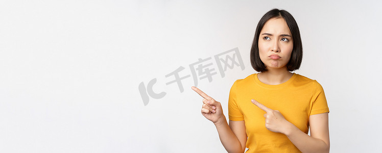脾气暴躁的亚洲女孩心烦意乱，噘嘴，看着不公平的东西，用手指指着横幅、标志品牌，站在白色背景的黄色 T 恤上