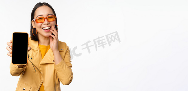 热情的亚洲女模特，在手机屏幕上展示智能手机应用程序界面、网上商店或网站，站在白色背景上